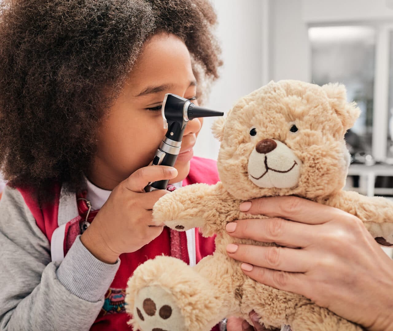 Young girl plays with her toy bear in a medical game, using an otoscope. Hearing clinic for children