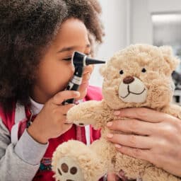 Young girl plays with her toy bear in a medical game, using an otoscope. Hearing clinic for children