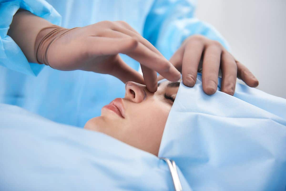 nasal-surgery-evaluation doctor's hands in rubber gloves are gently squeezing the bridge of a woman under a surgery cover with face exposed