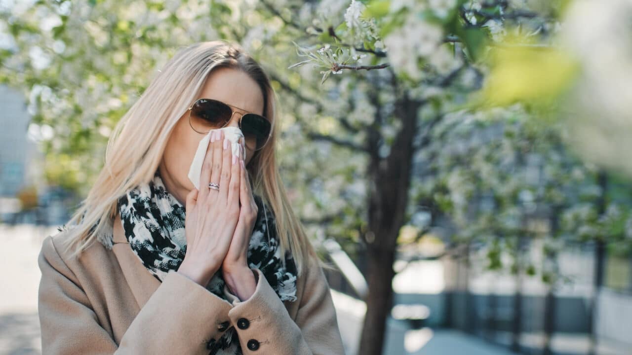 Woman blowing her nose near some trees.