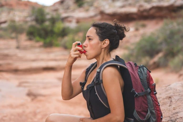 Asthma-inhaler Woman with a backpack on in a desert setting is using a red inhaler.