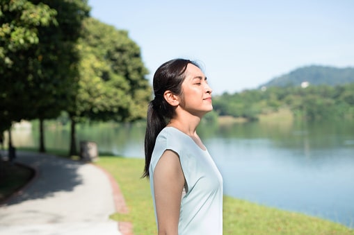 On a walk in a pleasant park by the lake, a woman stops for a deep, calming breath, unbothered by allergy symptoms. 